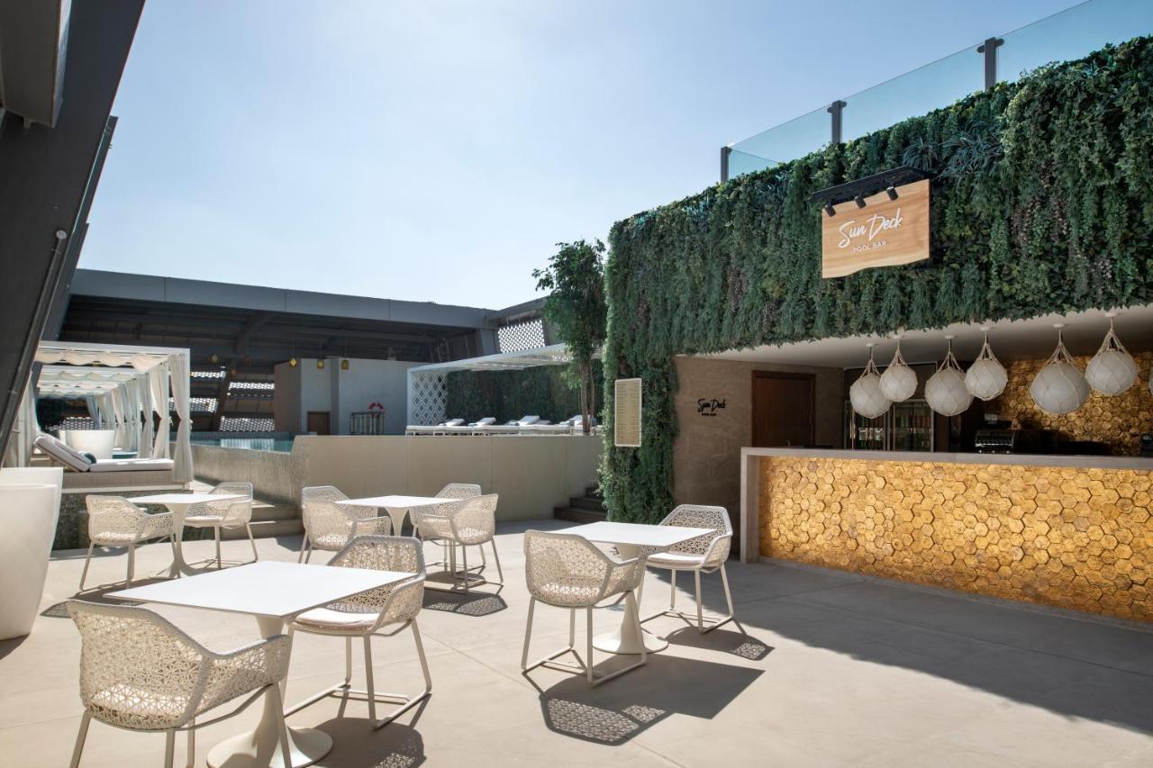 Steigenberger Hotel Doha Exterior foto The photo shows a stylish outdoor seating area of a modern venue, likely a restaurant or bar. The space features several white tables and chairs arranged neatly on a light-colored floor. In the background, there's a bar area with a warm-toned wood fa