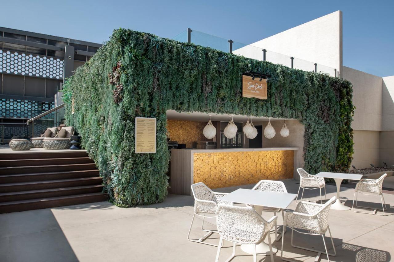 Steigenberger Hotel Doha Exterior foto The photo shows an outdoor bar area surrounded by lush greenery. The bar has a modern design, featuring a wooden counter with decorative pendant lights hanging above. There are several white outdoor tables and chairs arranged in front of the bar, whi