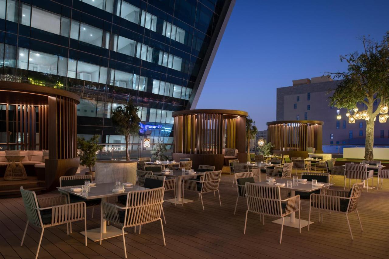 Steigenberger Hotel Doha Exterior foto The photo shows an outdoor dining area that features several tables and chairs arranged neatly on a wooden deck. The seating includes modern white chairs and a mix of seating styles, including some areas with privacy screens or structures that provid