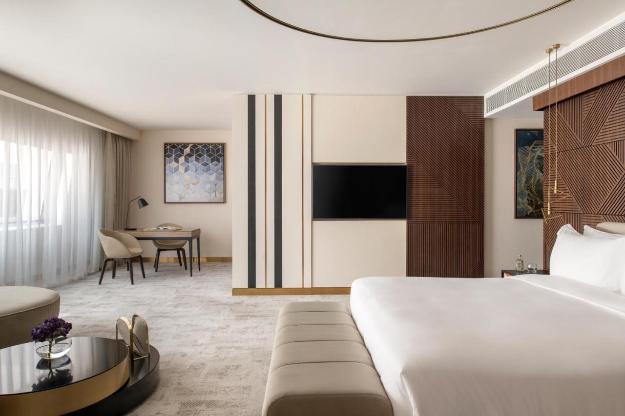 Steigenberger Hotel Doha Exterior foto The photo shows a modern hotel room with a contemporary design. It features a large bed with white linens, a stylish beige bench at the foot of the bed, and a sleek coffee table in front of a comfortable seating area. The room has a neutral color pal