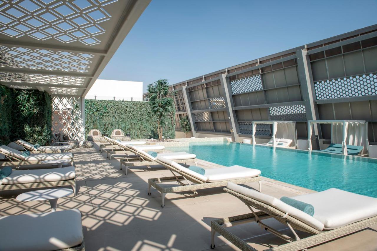 Steigenberger Hotel Doha Exterior foto The photo shows a modern outdoor pool area. There are several lounge chairs arranged around the pool, which is a vibrant blue color. Sunlight casts intricate shadows on the ground due to a lattice structure overhead. On one side, there are green plan
