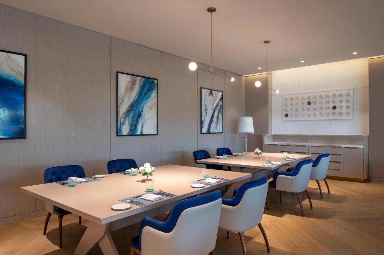 Steigenberger Hotel Doha Exterior foto The photo shows a modern, elegant dining or meeting room. It features a large, light-colored rectangular table surrounded by chairs with blue upholstery and white frames. Above the table, there are two hanging pendant lights providing illumination. T