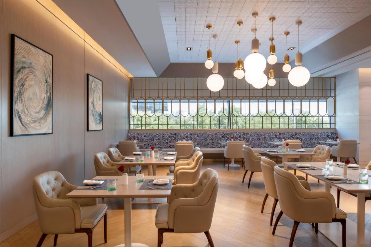 Steigenberger Hotel Doha Exterior foto The photo shows a modern restaurant interior featuring a clean and elegant design. There are several light-colored wooden tables with light-colored upholstered chairs arranged throughout the space. The walls are adorned with abstract art pieces, and 