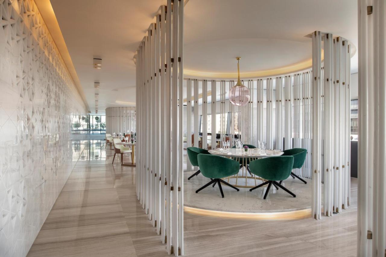 Steigenberger Hotel Doha Exterior foto The photo shows a modern, stylish interior of a restaurant or dining area. There is a circular dining table surrounded by green upholstered chairs with a sleek design. The space is framed by vertical white panels, creating a sense of separation and p