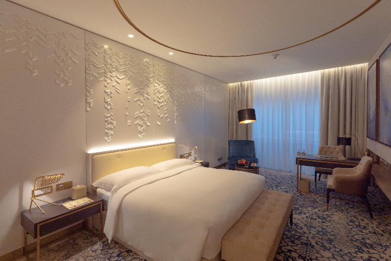 Steigenberger Hotel Doha Exterior foto The photo shows a modern hotel room featuring a contemporary design. The room includes a large bed with a padded headboard, positioned against a textured wall with decorative patterns. To the side, there is a stylish bedside table with a lamp. A comf